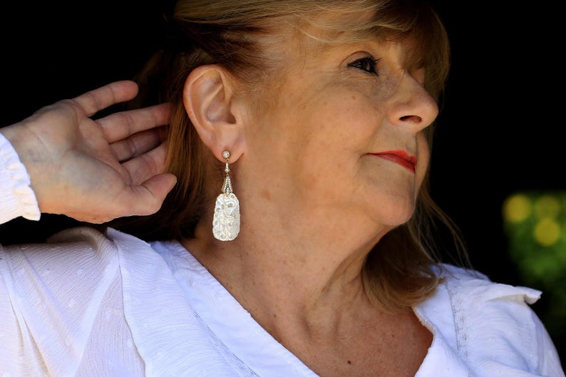 May include: A woman with blonde hair wearing a white shirt and silver dangle earrings with a floral design.