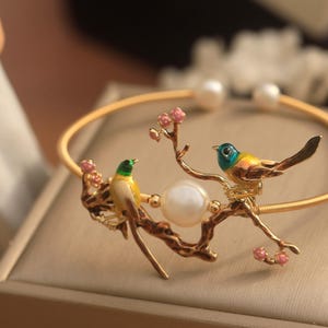 May include: A gold bracelet with a delicate design featuring two birds perched on a branch with pink blossoms. A pearl is incorporated into the design.