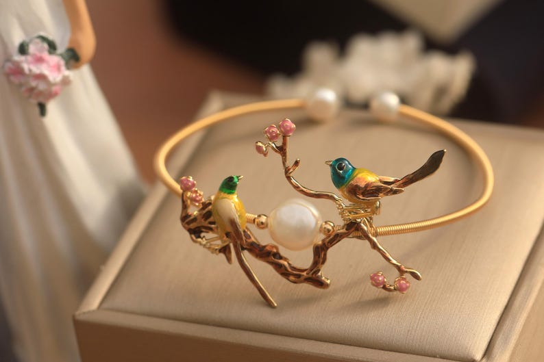 May include: A gold bracelet with a delicate design featuring two birds perched on a branch with pink blossoms. A pearl is incorporated into the design.