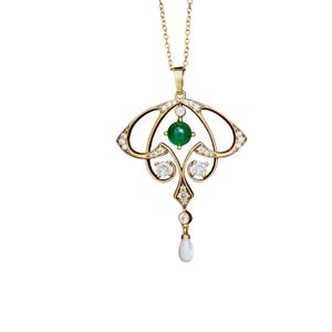 May include: A gold-colored pendant necklace featuring an Art Nouveau design. The pendant has a central green gemstone, surrounded by clear gemstones and a teardrop-shaped pearl. The necklace chain is also gold-colored.