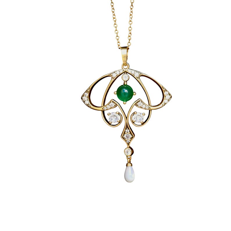 May include: A gold-colored pendant necklace featuring an Art Nouveau design. The pendant has a central green gemstone, surrounded by clear gemstones and a teardrop-shaped pearl. The necklace chain is also gold-colored.