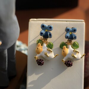 May include: A pair of gold earrings with a white and yellow ceramic mouse charm, a blue and yellow flower charm, and a dark purple berry charm.