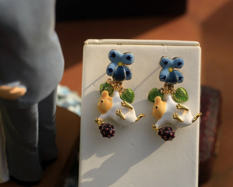 May include: A pair of gold earrings with a white and yellow ceramic mouse charm, a blue and yellow flower charm, and a dark purple berry charm.
