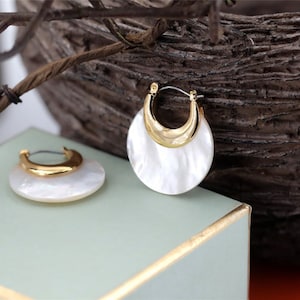 May include: A pair of gold hoop earrings with a white mother-of-pearl inlay. The earrings are round and have a slightly curved shape.