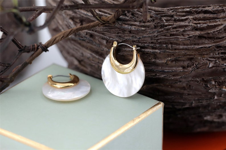 May include: A pair of gold hoop earrings with a white mother-of-pearl inlay. The earrings are round and have a slightly curved shape.