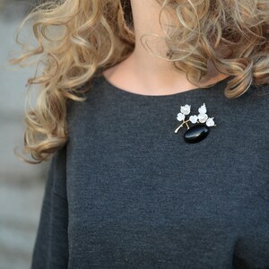 May include: A gold-toned brooch featuring a black oval stone and white floral accents. The brooch is attached to a gray top. The design includes a branch-like structure with delicate white flowers and pearls.