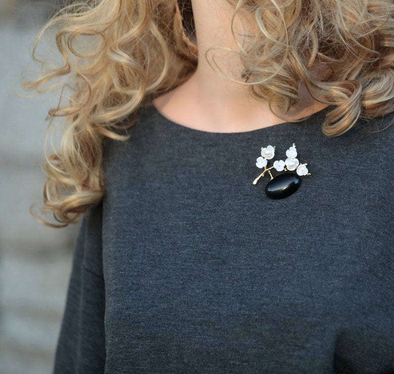 May include: A gold-toned brooch featuring a black oval stone and white floral accents. The brooch is attached to a gray top. The design includes a branch-like structure with delicate white flowers and pearls.