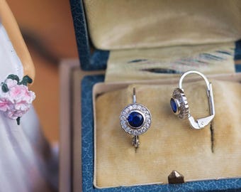 May include: A pair of silver earrings with a central blue gemstone surrounded by small clear stones. The earrings are displayed in an open blue jewelry box with a cream-colored interior.