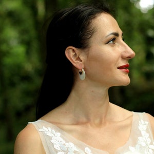 May include: A woman with long dark hair wearing a white dress with floral embroidery and silver hoop earrings. She is looking off to the side with a slight smile.
