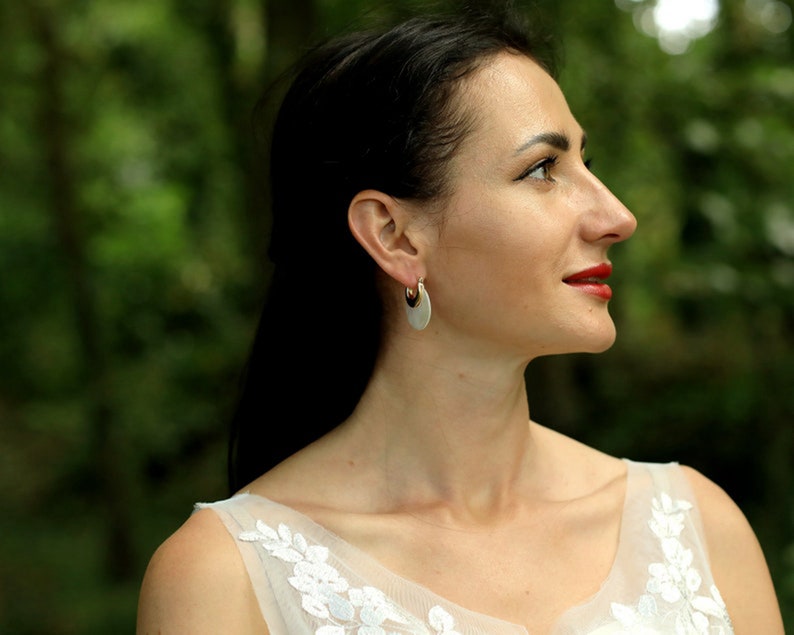 May include: A woman with long dark hair wearing a white dress with floral embroidery and silver hoop earrings. She is looking off to the side with a slight smile.