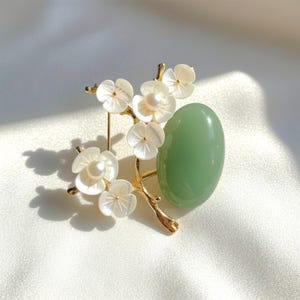 May include: A gold-toned brooch featuring a branch with white flower blossoms and a light green oval stone. The flowers have pearl centers. The brooch is set against a white, silky background.