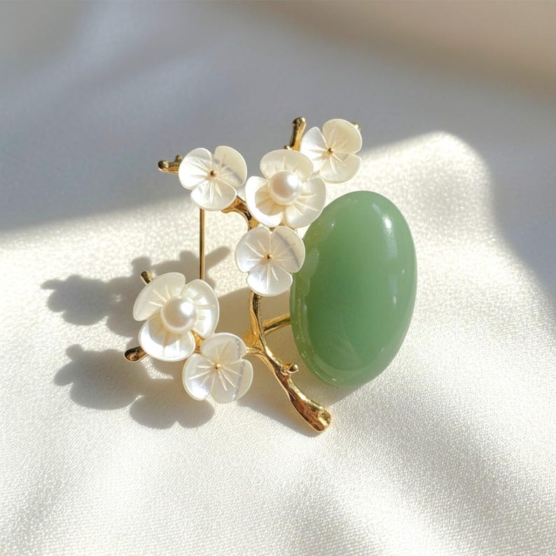 May include: A gold-toned brooch featuring a branch with white flower blossoms and a light green oval stone. The flowers have pearl centers. The brooch is set against a white, silky background.