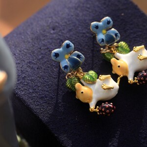 May include: A pair of dangle earrings featuring a white porcelain animal with a yellow head and blue flower accents. The animal is holding a small purple berry.