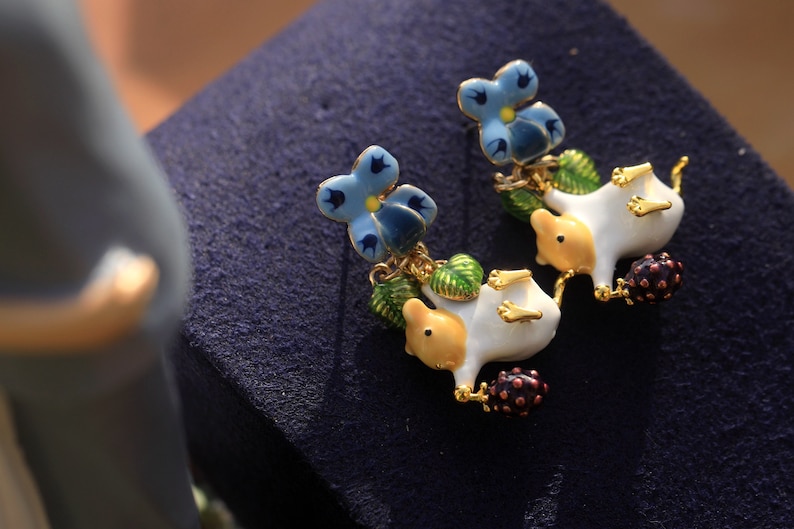 May include: A pair of dangle earrings featuring a white porcelain animal with a yellow head and blue flower accents. The animal is holding a small purple berry.