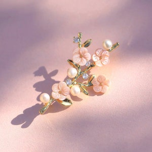 May include: A gold-tone brooch with a delicate branch design featuring pink flowers and pearl accents.