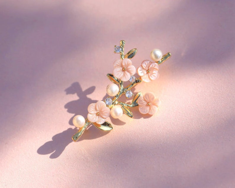 May include: A gold-tone brooch with a delicate branch design featuring pink flowers and pearl accents.