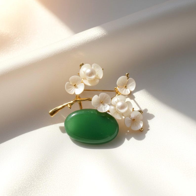 May include: A gold-toned brooch featuring a green oval stone and delicate white flower accents. The flowers have pearl centers and are arranged on a branch-like structure. The brooch is set against a white background.