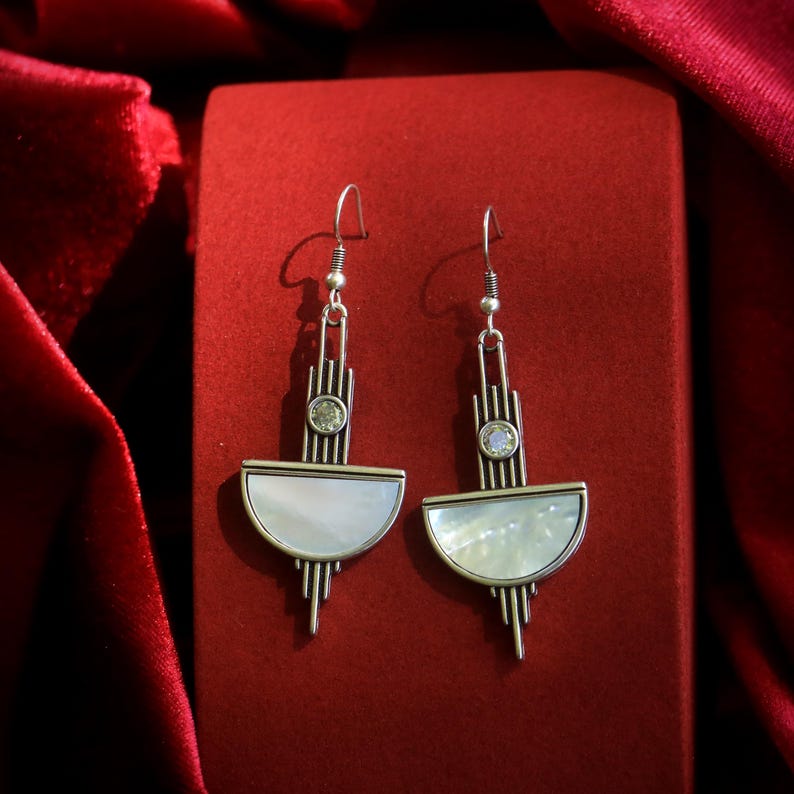 May include: A pair of silver-toned dangle earrings featuring a geometric design with vertical bars, a small circular clear stone, and a semi-circular white mother-of-pearl inlay. The earrings hang from simple silver-toned metal hooks.