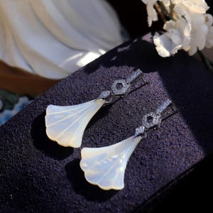 May include: A pair of elegant, fan-shaped earrings. Each earring features a pearly white, scalloped design, connected to a hexagonal, diamond-studded stud. The earrings are set against a dark purple background, with white flowers in the background.