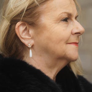 May include: A woman wearing a black fur coat with a pair of gold and white dangle earrings. The earrings feature a cluster of small white stones and a long, teardrop-shaped white stone.
