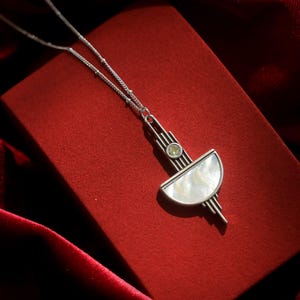 May include: A silver-toned necklace featuring a geometric pendant with a semi-circular mother-of-pearl inlay and a small, round, light green gemstone set above it. The pendant is suspended from a delicate silver chain with small, evenly spaced beads.