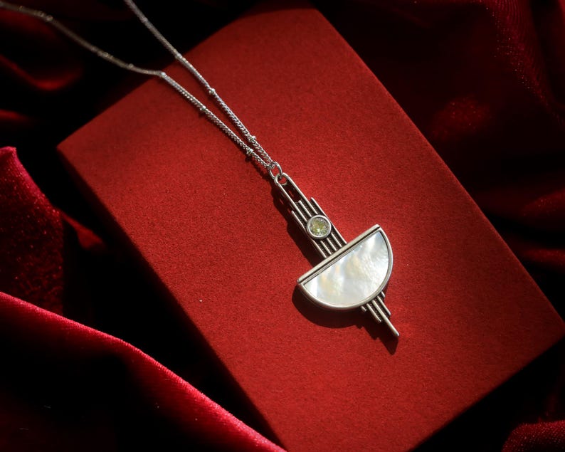 May include: A silver-toned necklace featuring a geometric pendant with a semi-circular mother-of-pearl inlay and a small, round, light green gemstone set above it. The pendant is suspended from a delicate silver chain with small, evenly spaced beads.