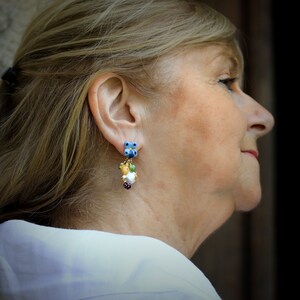 May include: A pair of dangle earrings with blue, green, white, and gold accents. The earrings feature a cluster of small charms, including a flower, a bird, and a berry.