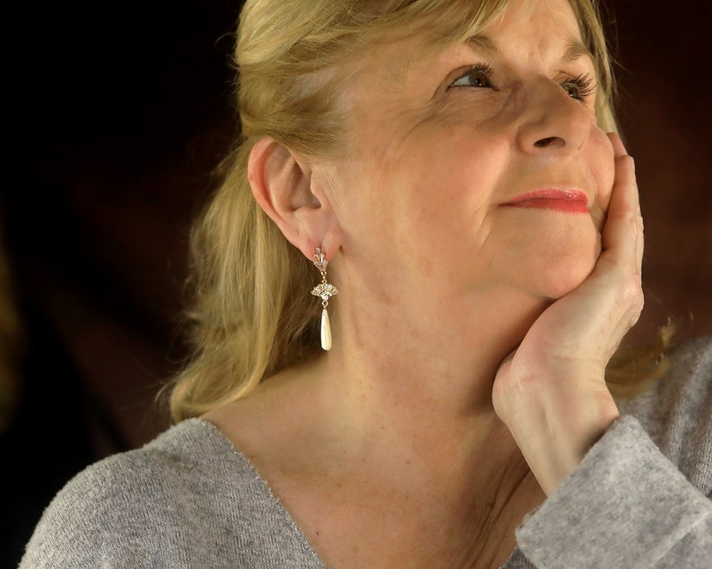 May include: A woman with blonde hair is wearing a pair of pearl drop earrings with a floral design. She is looking up and has her hand on her chin.