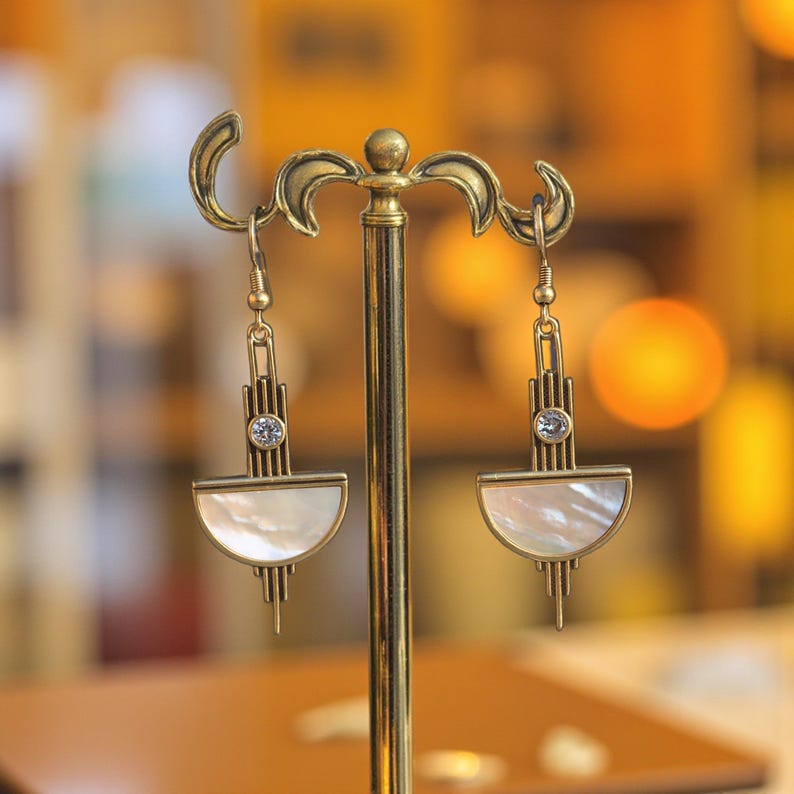 May include: A pair of antique brass-toned earrings hanging from a display stand. Each earring features a vertical bar design with a small clear round crystal and a semi-circular piece of iridescent white mother-of-pearl at the base.