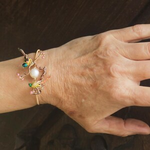 May include: A gold bracelet with a delicate design featuring a pearl, pink flowers, and two small birds with green and yellow accents.