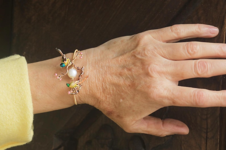 May include: A gold bracelet with a delicate design featuring a pearl, pink flowers, and two small birds with green and yellow accents.