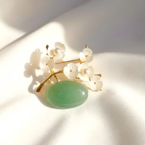 May include: A gold-toned brooch featuring a sprig of white flower blossoms with pearl accents and a large, oval, green stone. The brooch is set against a white, silky background.