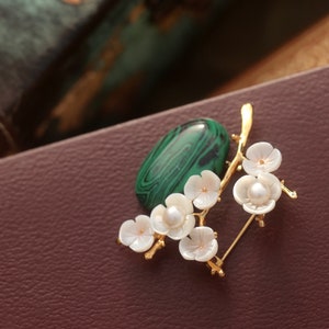 May include: A gold-toned brooch featuring a green malachite stone and a branch of white flower blossoms with pearl centers. The brooch is set against a brown leather surface.