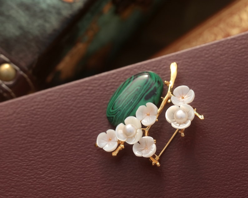 May include: A gold-toned brooch featuring a green malachite stone and a branch of white flower blossoms with pearl centers. The brooch is set against a brown leather surface.