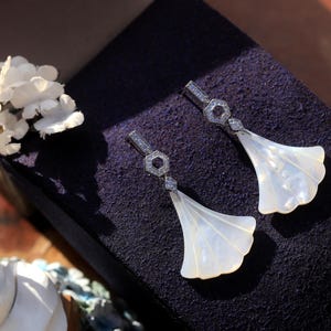 May include: A pair of elegant drop earrings featuring a fan-shaped, pearlescent white design. The earrings have a silver-colored, geometric top section with small, sparkling accents. The earrings are displayed on a dark purple surface, with white flowers in the background.