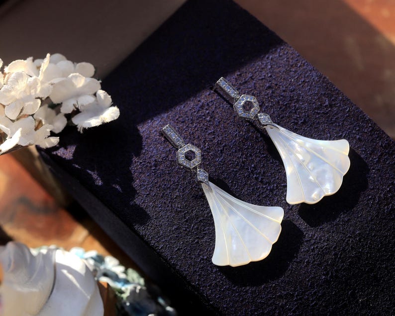 May include: A pair of elegant drop earrings featuring a fan-shaped, pearlescent white design. The earrings have a silver-colored, geometric top section with small, sparkling accents. The earrings are displayed on a dark purple surface, with white flowers in the background.