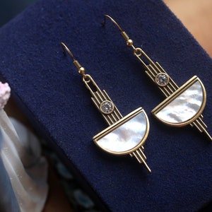 May include: A pair of gold-toned dangle earrings featuring a semi-circular mother-of-pearl inlay, a small clear round crystal accent, and a linear metal structure with vertical bars extending above and below the central design element.