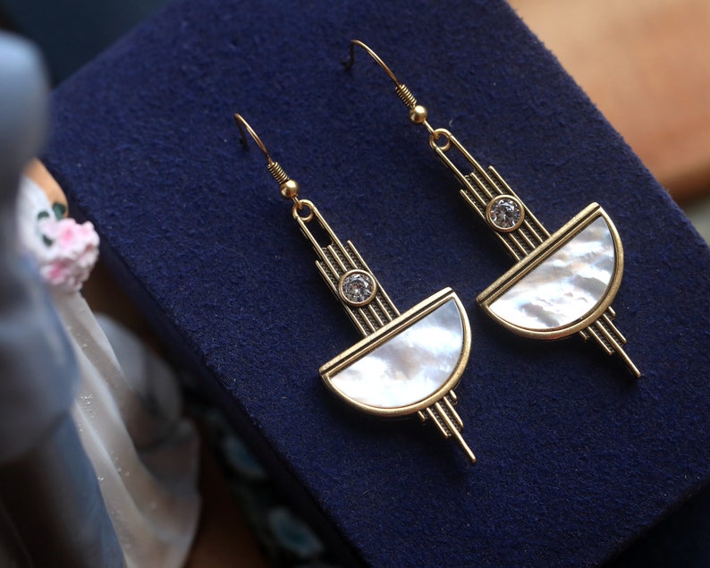 May include: A pair of gold-toned dangle earrings featuring a semi-circular mother-of-pearl inlay, a small clear round crystal accent, and a linear metal structure with vertical bars extending above and below the central design element.