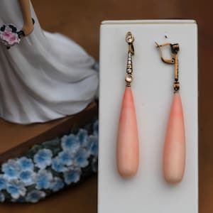 以下が含まれることがあります： 涙型のコーラルストーンと小さなダイヤモンドのアクセントが付いた、ドロップ型のイヤリングのペア。イヤリングは白いディスプレイカードの上に置かれています。