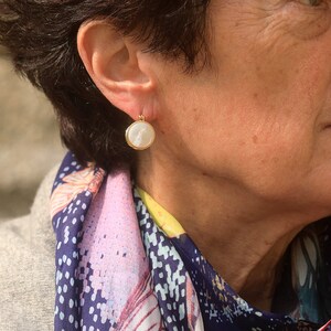 May include: A woman wearing a gold earring with a white pearl-like stone. The earring is round and has a gold rim. The woman is wearing a colorful scarf.