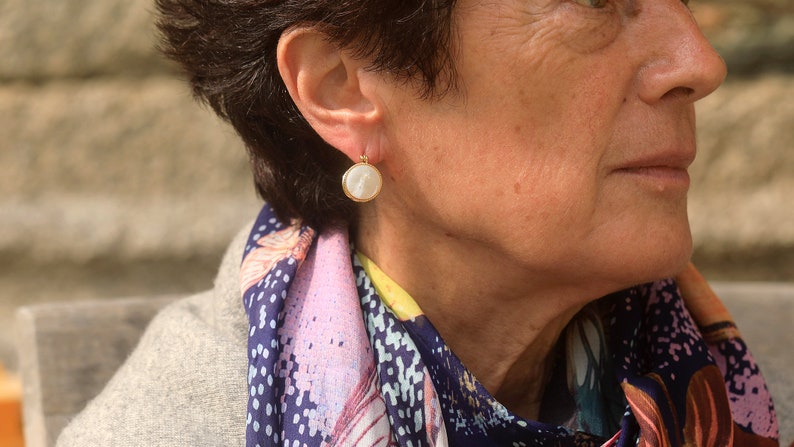 May include: A woman wearing a gold earring with a white pearl-like stone. The earring is round and has a gold rim. The woman is wearing a colorful scarf.