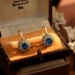 Puede incluir: Un par de pendientes de oro con una piedra preciosa azul rodeada de piedras blancas más pequeñas. Los pendientes están en una caja de terciopelo.