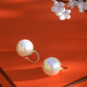 May include: A pair of gold hoop earrings with large, white pearl-like accents. The earrings are on a bright orange surface.