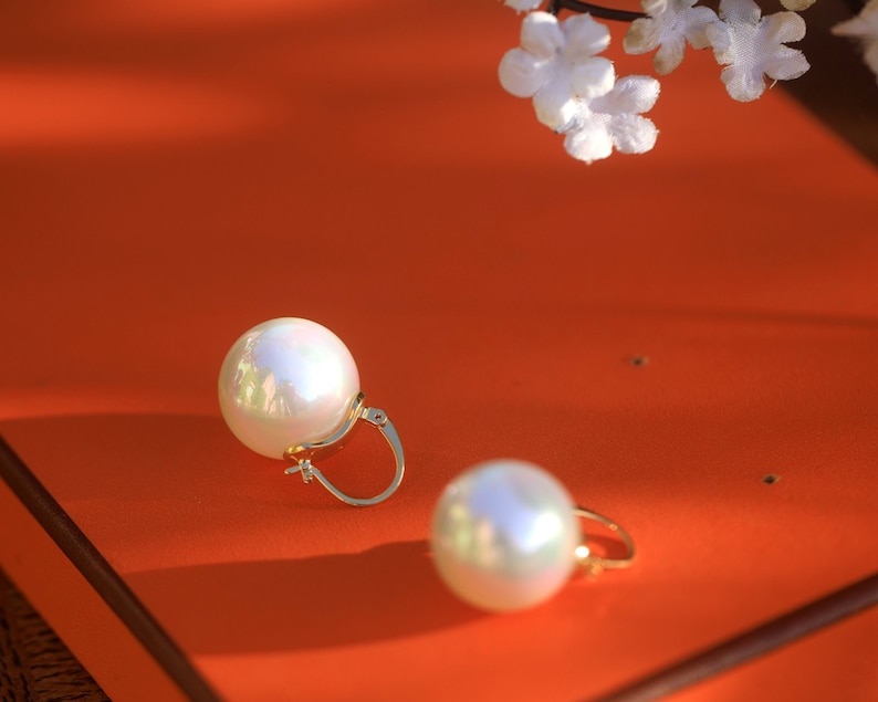 May include: A pair of gold hoop earrings with large, white pearl-like accents. The earrings are on a bright orange surface.