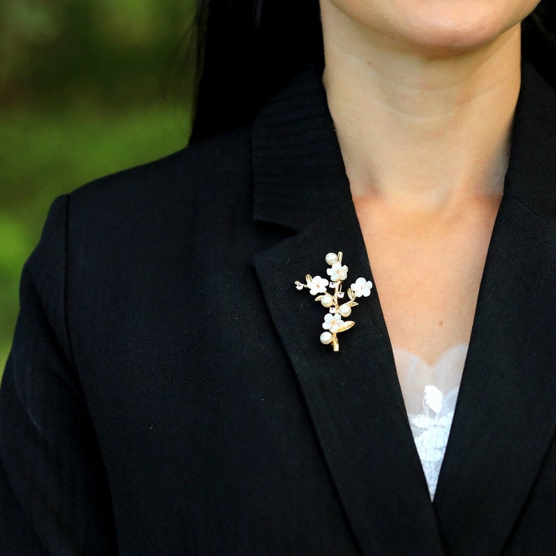 May include: A black blazer with a gold and white floral brooch pinned to the lapel.