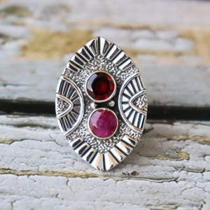 May include: A silver ring with a geometric design and two red gemstones. The ring is oval shaped and has a detailed pattern.