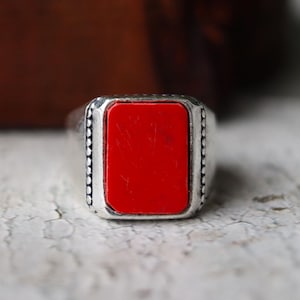 Könnte beinhalten: Silberring mit einem rechteckigen, leuchtend roten Stein. Der Ring hat einen perlenbesetzten Rand um den roten Stein. Der Ring ist vor einem neutralen Hintergrund platziert.