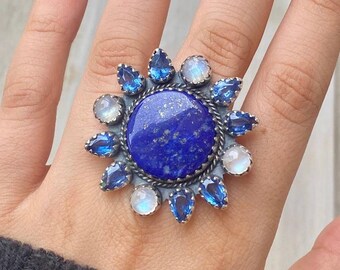 May include: A large, ornate ring featuring a deep blue, round gemstone at its center, surrounded by smaller blue and white stones. The ring has a silver-colored metal setting, creating a striking contrast with the vibrant gemstones. The design resembles a sunburst.