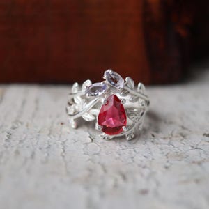 Puede incluir: Un anillo plateado con una gran gema roja en forma de pera como punto focal. El anillo está adornado con gemas moradas ovaladas más pequeñas y delicados detalles en forma de hojas. El diseño del anillo es intrincado y de inspiración natural.