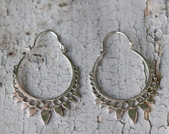 May include: A pair of silver hoop earrings. Each earring has a scalloped design along the bottom edge, with leaf-shaped pendants. The earrings are displayed on a weathered, grey surface.